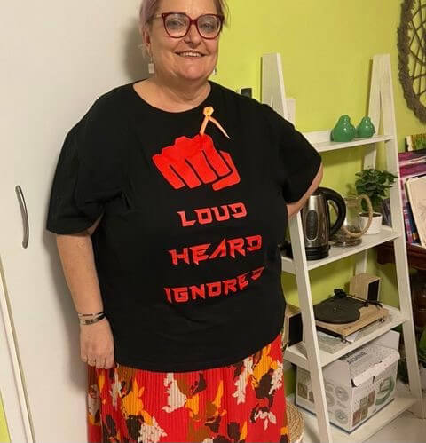 Laura with one hand on her hip, standing against a white door, smiling at camera. She has a short pink pixie cut, wears red glasses and a multicoloured skirt and black top with a cartoon of a red closed fist and the words "Loud. Heard. Ignored."