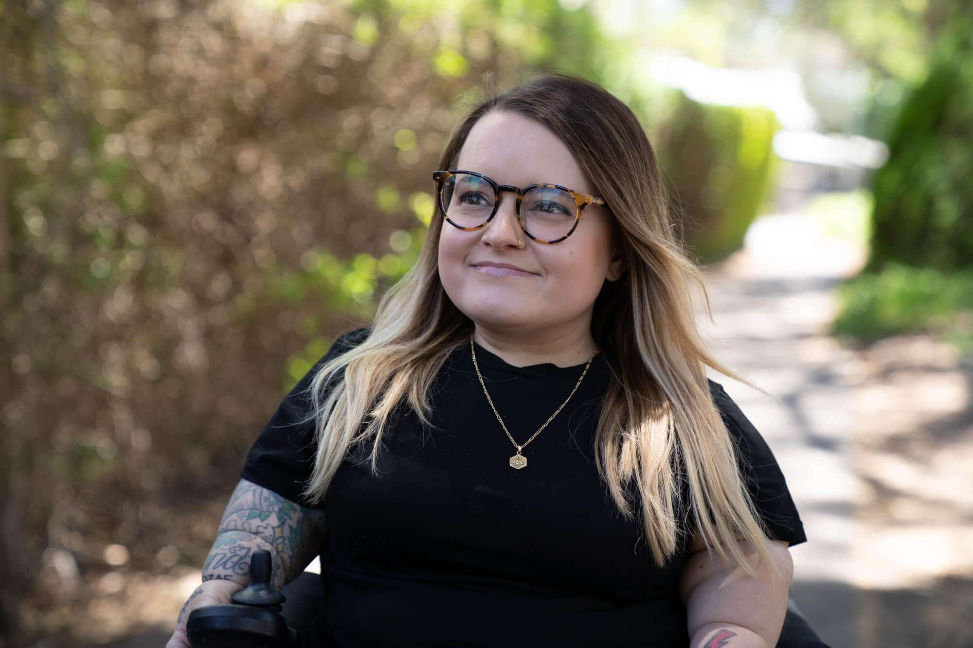 Photo of Belle in her motorised wheelchair outdoors. She wears a short sleeved black shirt which shows her tattooed sleeve. She has tortoiseshell framed glasses and dark brown hair with blonde tips down to her shoulders, and is smiling.