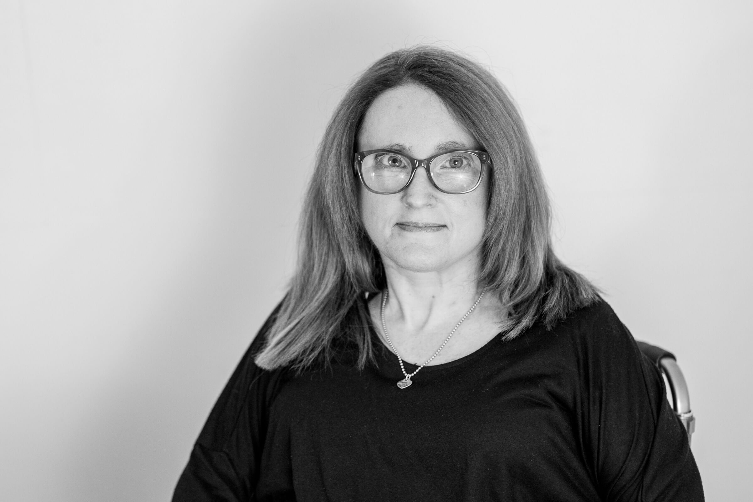 A black and white photo of Ainslee looking at the camera. She has shoulder length straight hair framing her face, wears glasses and a necklace, and a dark coloured top.