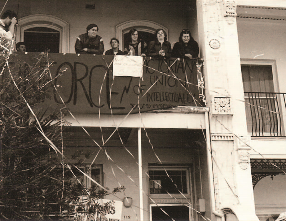 Image is of Reinforce protesters on the top balcony of 112 Drummond Street with a sign that says: "Reinforce: Victorian Union of Intellectual Disability"