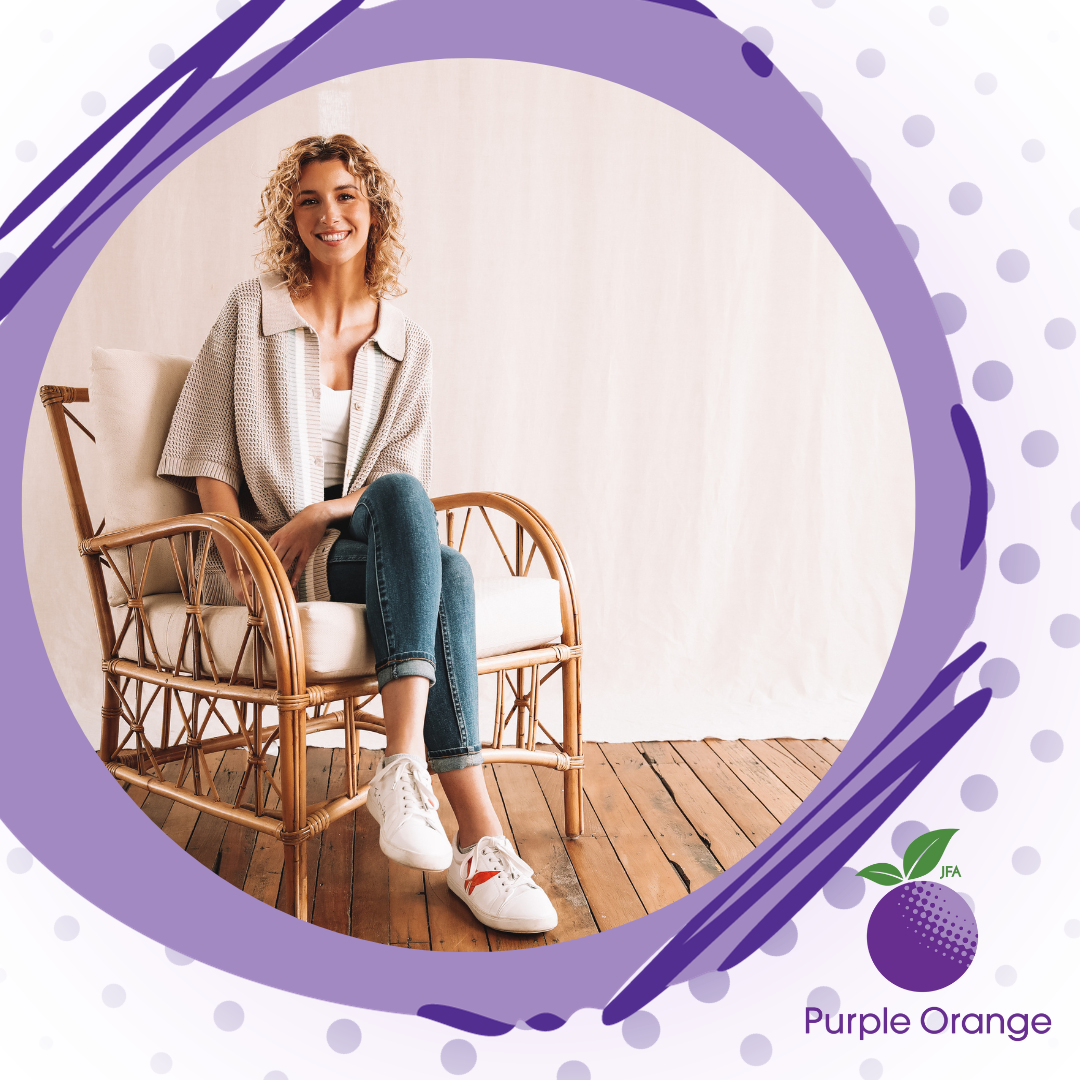 Light background with purple dots, dusty and dark purple circular accents around photo of Lauren, sitting in a wicker chair, wearing jeans and light colours. Lauren and shoulder length curly hair and smiles at the camera.