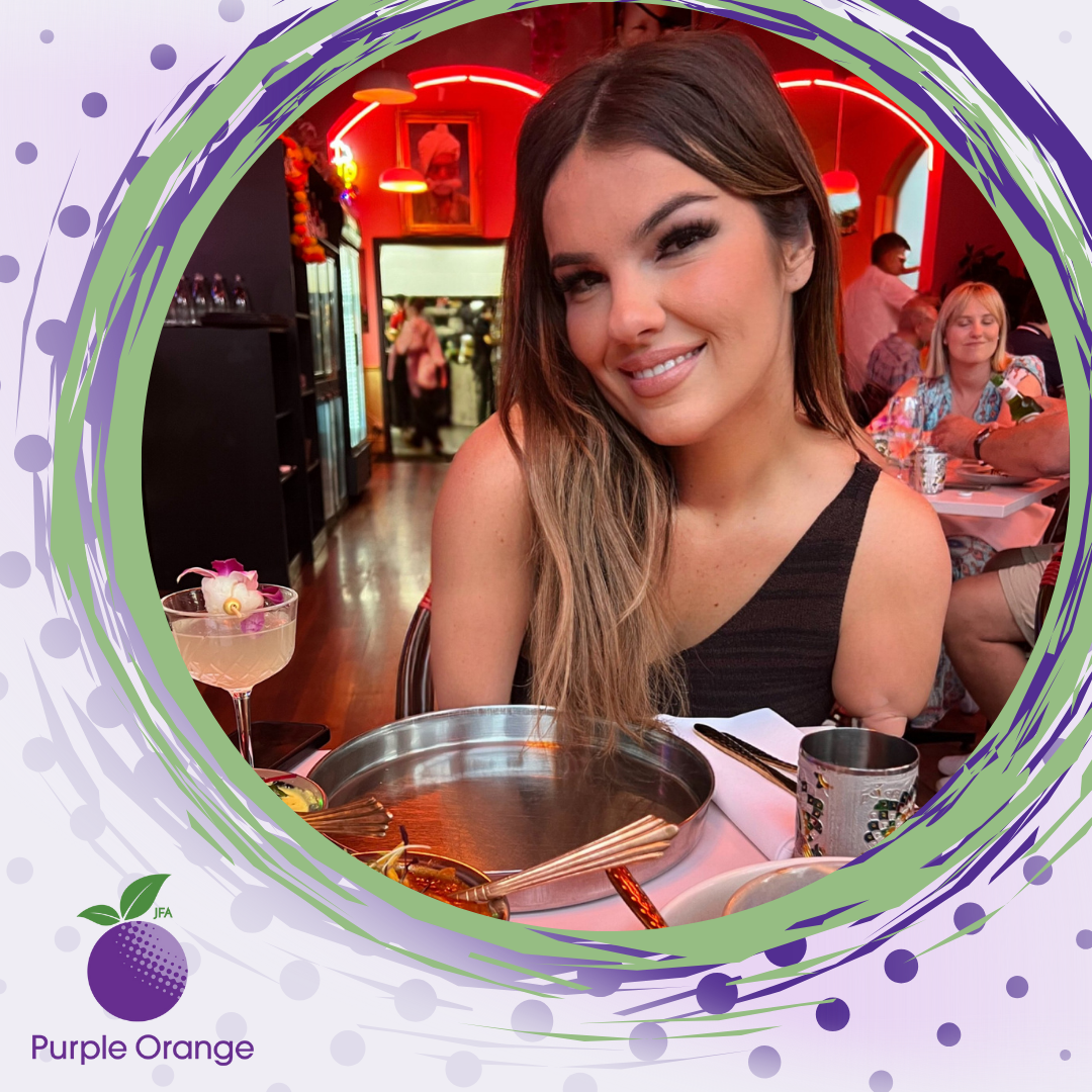 a light purple spotted background with a photo of Daniella at a resturant, dressed up, smiling at the camera. Plates of food in front and fancy lighting behind.