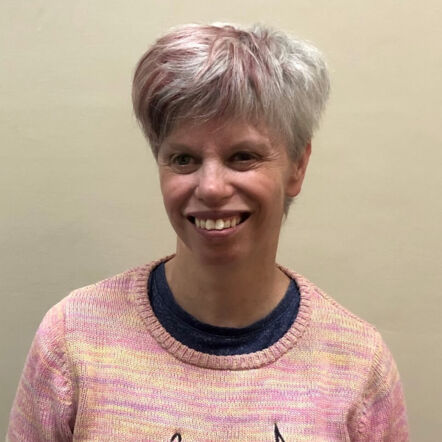 Photo of Shannon who has short grey hair with light pink streaks, wearing a pink jumper and smiling off camera.