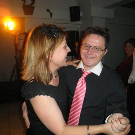 Photo of Louise at her brother's 50th birthday party. She wears a black top with a black fascinator in her light brown shoulder length hair. She is dancing hand in hand with her brother.