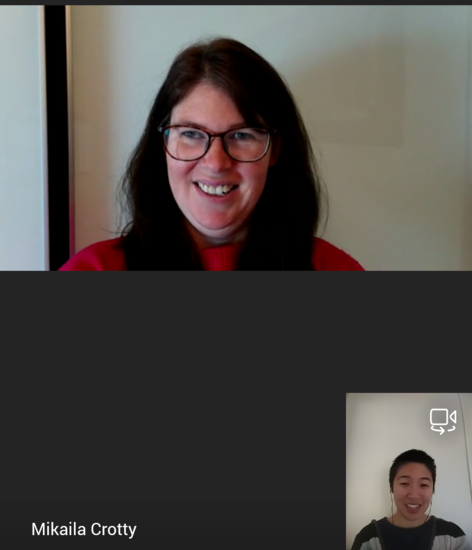 A Microsoft Teams call between Mik and Isabel. Mik is wearing glasses and a red top. Isabel is in a grey top with a striped jumper over her top and wearing earphones. Both are smiling at the camera.