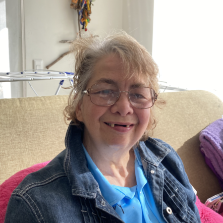 Photo of Maria sitting on a beige sofa. She is an elderly Aboriginal woman with short white hair, wearing glasses and a collared denim jacket over a bright blue top, grinning happily at the camera.