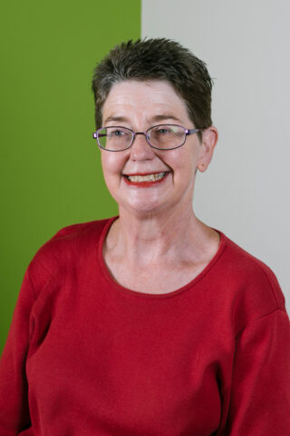 Photo of Annette smiling at something off-camera. She is a white woman with glasses and short dark hair, wearing a red long sleeved shirt, standing against a split green and grey background. 