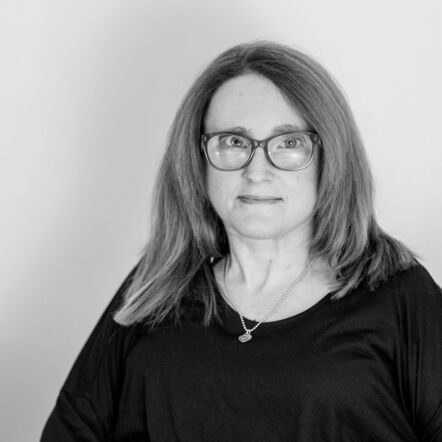 A black and white photo of Ainslee looking at the camera. She has shoulder length straight hair framing her face, wears glasses and a necklace, and a dark coloured top.