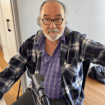 Photo of Marcelo looking at camera. He's an older man with grey hair and olive skin, wearing glasses, a grey plaid jumper over a purple collared shirt.