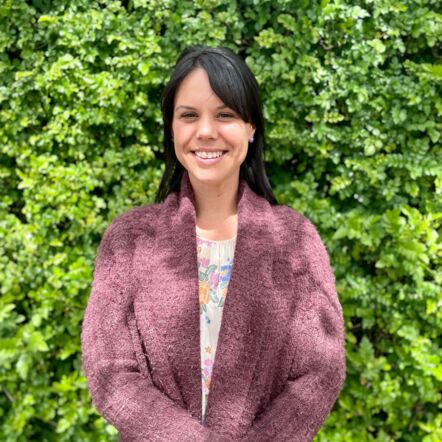 Photo of Ayla in front of green wall, smiling at camera. She has olive skin, dark hair parted to the side, and wears a patterned blouse with a plum coloured jumper over the top.