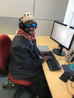 Dr Siyat takes a break from his computer at work to smile for the camera. He is wearing dark tinted glasses, dressed in a smart black blazer and wearing a scarf around his neck.
