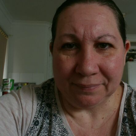 A selfie of Donna, with the background of a kitchen. She is a woman with dark hair tied back, wears a grey top with a black swirl pattern in the front, and is looking straight into the camera.