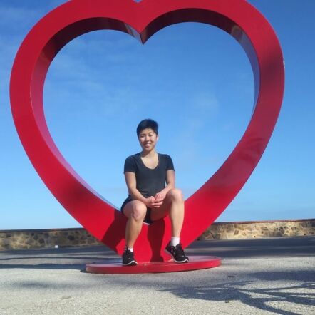 Photo of Isabel, who has dark short hair in a pixie cut, sitting on a red heart sculpture at Glenelg beach.