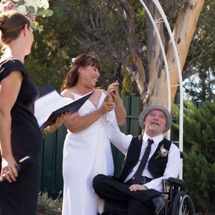A Photo of Stephen, who is blind and uses a wheelchair and his wife Tracy under the wedding altar. She is in a wedding dress. He is in a suit. They are smiling and holding hands.