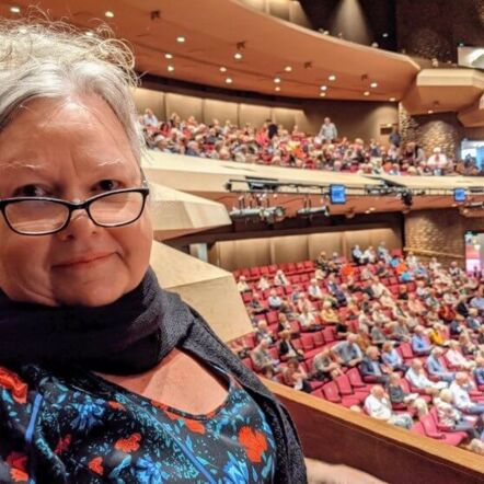 Photo of Julie indoors in a crowded amphitheatre. She has grey hair that's swept back, black framed glasses and wears a black top with a floral pattern and a black scarf around her neck.