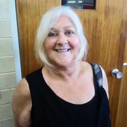 Photo of Annie beaming at the camera in front of a wooden door. She is a woman with blue eyes and light blonde hair up in a ponytail, and wears a black sleeveless top. 