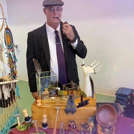 Photo of Martin in a circle frame on a dark purple background. Martin is standing behind a table of wooden items that he has produced, including a bowl, rings, figurines, a dream catcher, shaving brushes and more. Martin wears a dark suit, white shirt and tie and a bowling cap. He has a pipe in his mouth. 