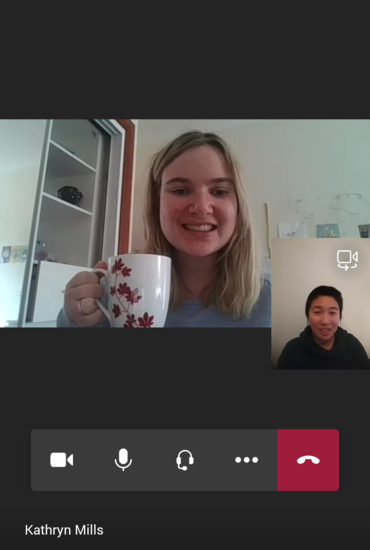 Teams call between Kathryn and Isabel. Kathryn is in a light blue top and holding up her white patterned mug. Isabel is in a dark green jumper with earphones in. Both are smiling at the camera.