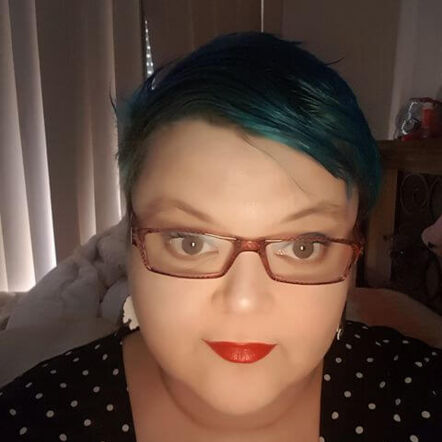 A selfie of Eva, who has short cropped turquoise hair, wears red lipstick and glasses with red frame.