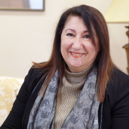 A photo of a woman with dark shoulder length hair, smiling at the camera.She is wearing a khaki turtleneck top and black blazer, with a grey scarf with a pattern of elephants around her neck.