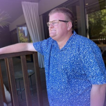 Photo of Cameron pictured slightly from the side, standing, with his arm resting on a high chair arm. He is wearing glasses and a blue patterned shirt.