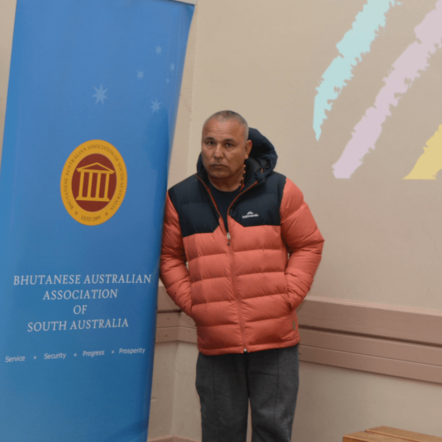 Krishna, an older man with brown skin, stands next to the Bhutanese Community banner.