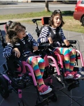 A photo of Fatimeh's daughters, Hediyeh and Haniyeh. Both girls are dressed in matching outfits, a long sleeved striped top and rainbow leggings. They are in their wheelchairs and looking off camera with big smiles.