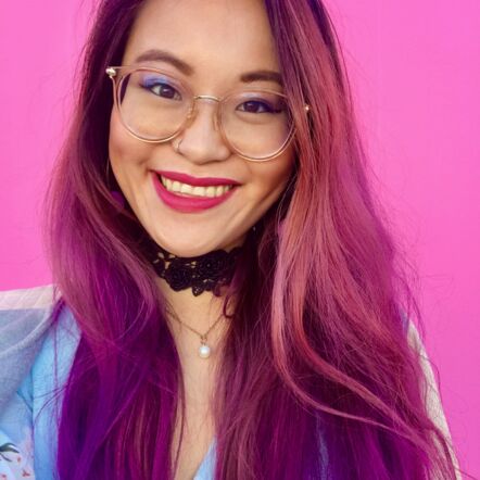 Photo of Akii smiling at the camera. They are Asian, wear glasses and have long bright pink hair with purple streaks at the ends.