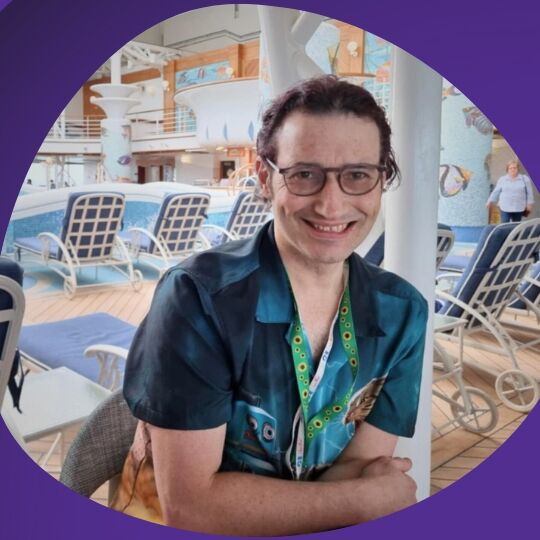 Photo of a smiling Daniel seated at a table with sun loungers in the background. He is wearing a patterned, blue short sleeve shirt. He has short dark hair and wears black glasses. His photo is in an organic round shape with a purple background behind it.