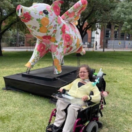 Alice sits in her wheelchair outdoors on the grass. She wears a yellow top and glasses and is smiling. Behind her is a large art sculpture of a pig with wings, a pattern of red and orange flowers across its body.