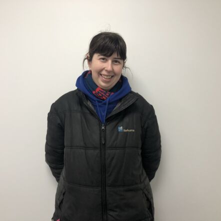 Photo of Elizabeth smiling at the camera and standing in front of a white wall. She has long black hair that is tied back, and wears a navy scarf with red stripes around her neck and a black jacket.