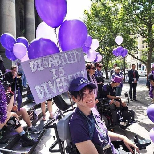 Image is a photo of Tiffany, dressed in purple with a big purple sign that says 'Disability is Diversity.' She is amongst a crowd of people also wearing purple and with lots of purple balloons.