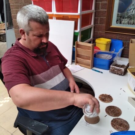 Photo of Daniel, a man with grey hair wearing a maroon top, sticking broken twigs to wooden coasters at his desk.