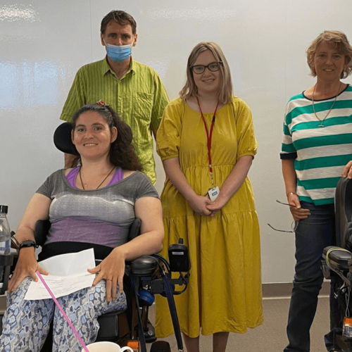 Photo of DEAA group members smiling at the camera. From left to right, Jill, Tiffany and Jane are in the front row in their wheelchairs. Behind them, standing, are Anthony who has a mask on, Kathryn, Jane and Megan.
