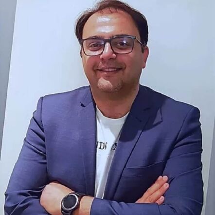 Photo of John, a man with dark hair and brown skin dressed in a navy blazer over a white shirt. A pair of glasses frame his face, and he is flashing a smile at the camera, with arms crossed in front of his chest.