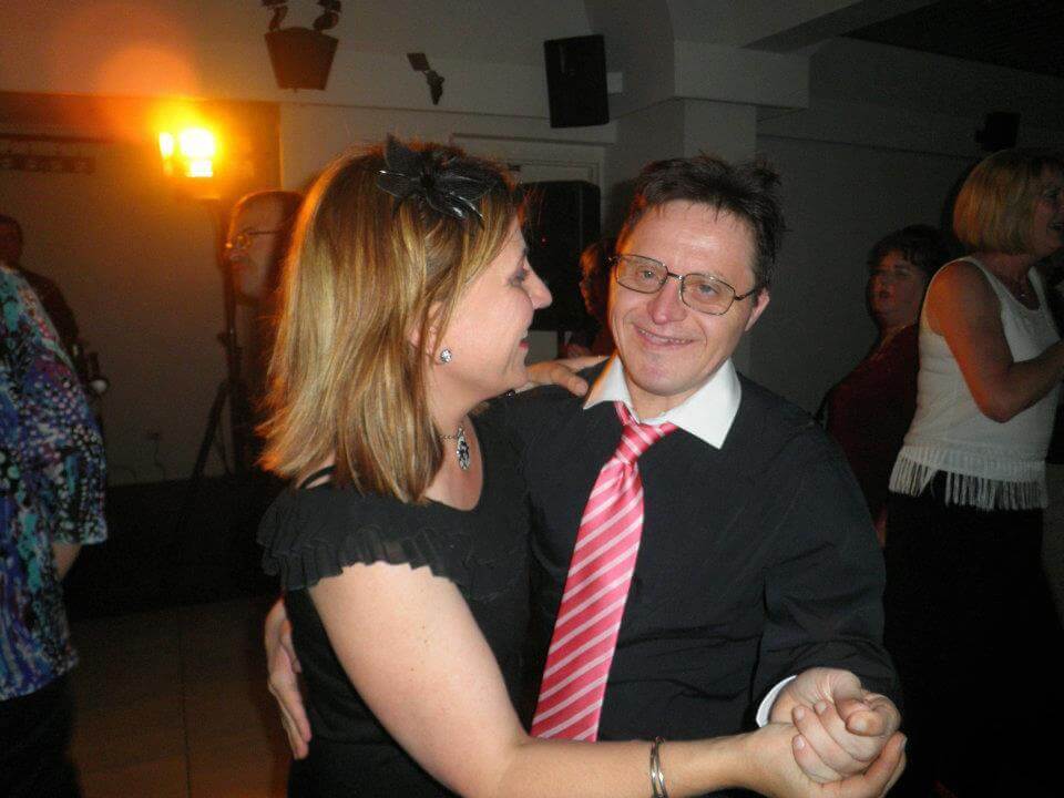 Photo of Louise at her brother's 50th birthday party. She wears a black top with a black fascinator in her light brown shoulder length hair. She is dancing hand in hand with her brother.