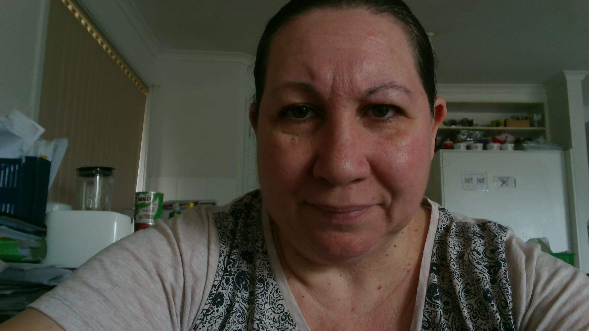 A selfie of Donna, with the background of a kitchen. She is a woman with dark hair tied back, wears a grey top with a black swirl pattern in the front, and is looking straight into the camera.