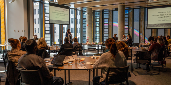 Photo of presenter speaking to a room of people sitting at circular tables Photo of presenter speaking to a room of people sitting at circular tables