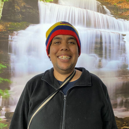 Photo of Carissa standing in front of a picture of a waterfall. She is an Aboriginal woman with a colourful beanie over her dark hair, wearing a black jumper and flashing a big smile at the camera.