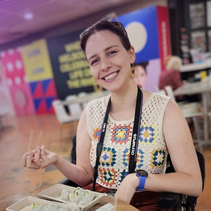 Photo of Tessa sitting at a café table, smiling in a sleeveless, crocheted top. She holds chopsticks in one hand. 