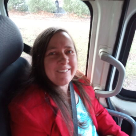Photo of Susan smiling at camera, from the seat of a vehicle. She has long dark shoulder length hair and wears a blue top with a bright red blazer.