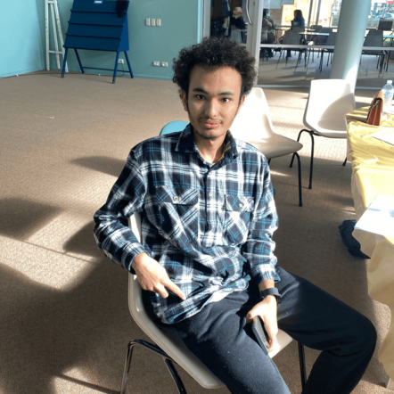 Photo of Prabin, a young man with dark curly hair and brown skin, sitting on a chair and smiling at the camera.