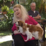 Photo of Ann Marie Smith in a garden. She has long light blonde hair down to her shoulders and is holding a dog in her lap, smiling.
