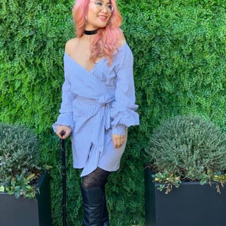 Photo of Akii standing in front of a green leafy wall, with two potted plants on either side. They are Asian and have long pink hair, wear glasses and are dressed in a light blue off-shoulder long sleeved dress with black tights. They hold a walking stick and are smiling to something off camera.