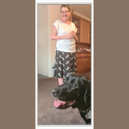Graphic of Polaroid photo showing Annette indoors holding a glass in her hands, looking down and smiling at her guide dog Molly, a black Labrador. Annette is a woman with white skin and short cropped hair, wears glasses and is dressed in a white top and dark grey patterned skirt. Molly is photobombing the shot with tongue out. Background of wooden kitchen cupboards and white walls.