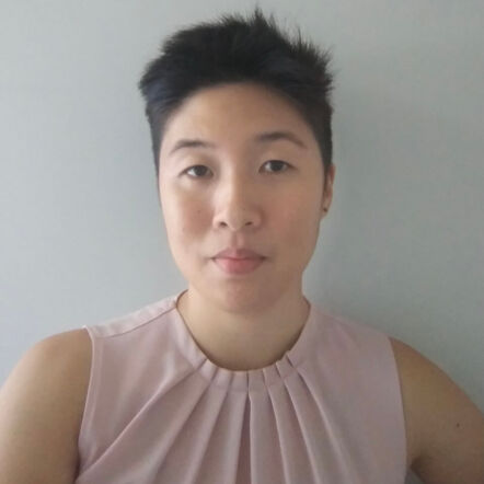 Selfie of Isabel against a grey background. She is an Asian woman with short black cropped hair and wears a sleeveless pink blouse.