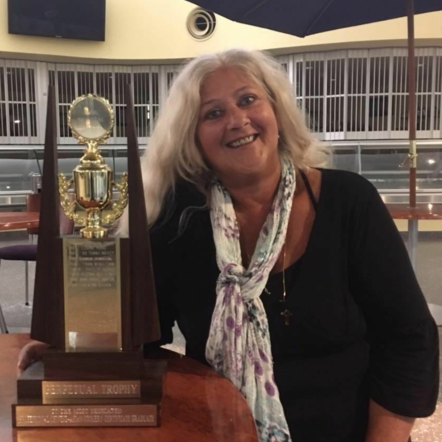 Photo shows Annie, a woman with tanned skin and shoulder length grey hair, who wears a black long sleeved top and a patterned white scarf around her neck. She is standing next to a large trophy which she won as the "Dedicated Chef - Chinese Asian Chef " award.