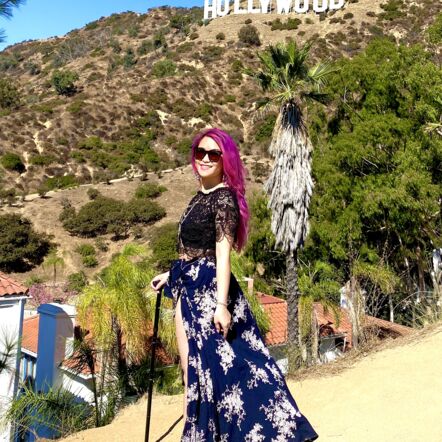 Photo of Akii in front of the Hollywood sign. They have long, bright pink hair, wear tinted sunglasses and hold a black cane.