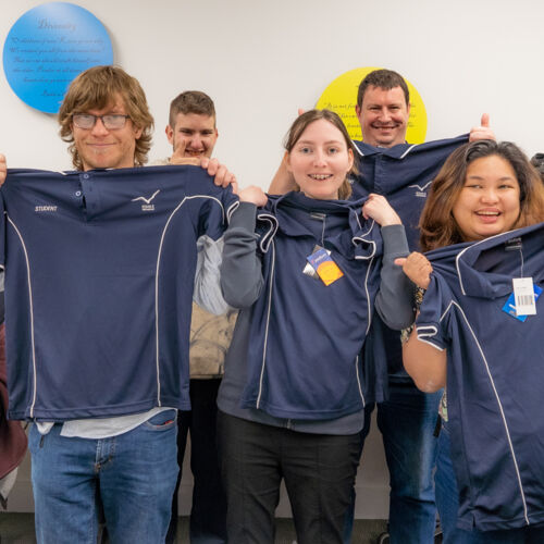 A mixed gender group of Aged Care trainees holding up their work shirts.
