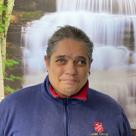 Photo of Sue standing in front of a picture of a waterfall. She is an Aboriginal woman with short grey hair, wearing a navy coloured Salvation Army Home League jumper.